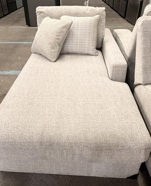 A light beige textured fabric chaise lounge chair with two matching accent pillows, shown in a showroom setting.